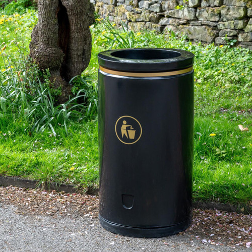 Roadware Coniston 90 Litre Open Top Litter Bin, black outdoor waste bin placed on a sidewalk with green grass, trees, and stone wall in the background.