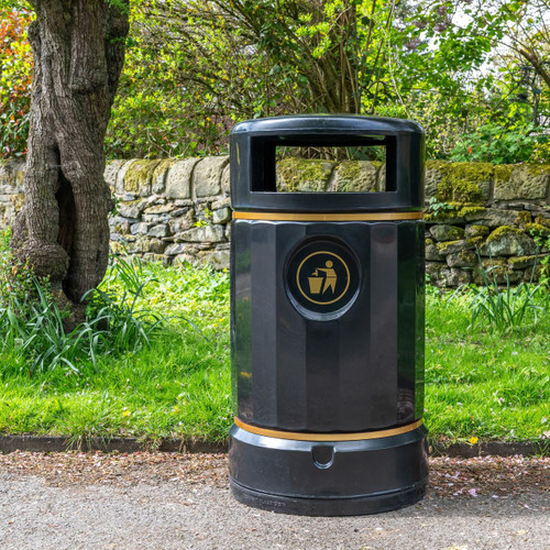 Roadware Derwent 120 Litre Classic Heritage Litter Bin, outdoor waste disposal container placed on a sidewalk near a grassy area with trees and a stone wall in the background.