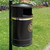 Melba Swintex Nickleby Heritage Post / Wall Litter Bin 35 Litre, black waste bin with a gold logo and a lid, mounted on a pole outdoors on a grassy area.