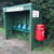 Melba Swintex Osprey Post / Wall Mount Litter Bin 35 Litre, green outdoor litter bin with seating shelter, mounted on a wooden structure, equipped with a red waste bin attached on the side.
