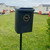 Oaklands 25L Apollo Post / Wall Dog Waste Bin, black mounted dog waste bin with a sticker that reads 'Dog Waste Accepted in This Bin' and a silhouette of a dog, placed outdoors in a grassy area with small yellow flowers and houses in the background.