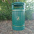 Oaklands Centurion Heritage Outdoor Litter Bin 90 litre, green metal outdoor trash bin with a recycling symbol on the front, placed on grass near bushes.