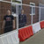 Oaklands Mesh Hoarding Panel For Sitewall Barrier, image showing white metal mesh panels attached to red and white plastic barriers with two people standing behind the panels