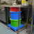Excelsior 70 Litre Inter Stacking Food Grade Plastic Ingredients Bin, colorful plastic stacking bins in red, blue, and green on a blue base under stainless steel worktable in a commercial kitchen.