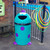 Leafield Monster Buddy School Litter Bin 100 Litre, colourful cartoon-inspired outdoor trash bin with a smiling face, purple feet, and purple spots, positioned next to a black metal fence with colorful hoops, in a school playground setting.