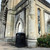 Leafield 240 Litre XL Square Heritage Outdoor Litter Bin, black outdoor litter bin placed next to a bench outside a historic stone building.