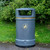 Roadware Derwent 120 Litre Outdoor Heritage Litter Bin, grey outdoor trash can with a large opening and a gold band, located on a grassy area with green bushes in the background.