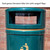 Roadware Derwent 120 Litre School Playground Litter Bin, dark green trash bin with black seagull flaps and a gold detail on the front, placed against a brick wall.