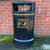Roadware 120L Derwent Large Floor Mount Dog Waste Bin, black durable outdoor waste bin with gold lettering and icons for litter and dog waste, placed against a brick wall on the pavement.