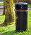 Roadware Coniston 90 Litre Outdoor Litter Bin, black cylindrical outdoor trash can with a gold accent line and a symbol of a person throwing trash, placed outdoors near a tree on grass.