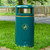 Roadware Coniston 90 Litre Outdoor Litter Bin, green outdoor trash can with a gold icon of a person disposing of litter, placed on grass near a park pathway.