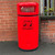 Roadware 90L Coniston Floor Mount Dog Waste Bin, large red outdoor litter and dog waste disposal bin with clear top flap and black base, placed against a brick wall.