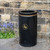 Roadware Coniston 90 Litre Open Top Litter Bin, black outdoor trash receptacle with a gold-colored symbol of a person disposing of rubbish, placed outdoors against a stone wall with purple flowers nearby.