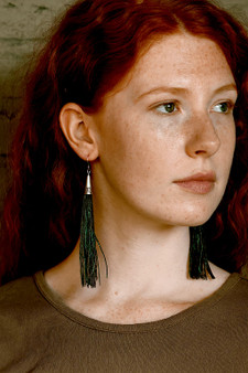 A woman wearing long tassel-style feather earrings made from iridescent green peacock herl with silver cone tops.