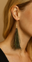 Close-up of a long tassel-style feather earring made from shimmering green peacock herl feathers, finished with a polished gold cone bead. The earring hangs gracefully against the wearer’s neck, showing fine iridescent detail.