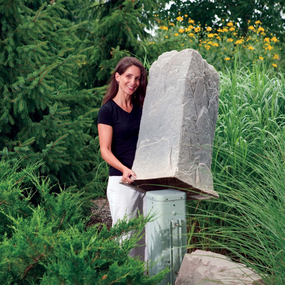 A woman lifting the lightweight DekoRRa Model 113 rock cover to conceal a tall utility pedestal, demonstrating its ease of use and natural appearance.