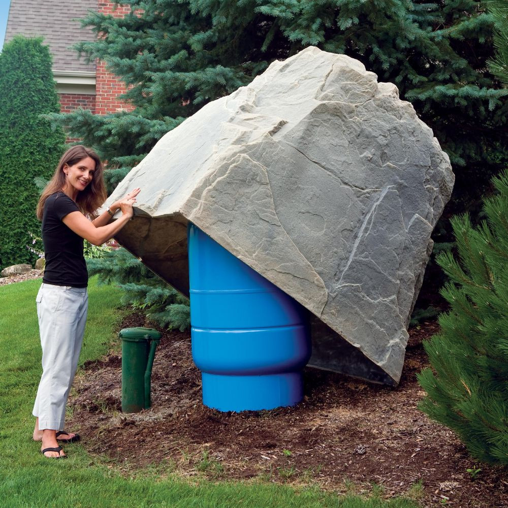 A woman lifting the DekoRRa Model 114 rock cover to conceal a large utility or septic component, demonstrating its lightweight yet durable construction.
