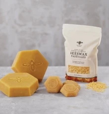 A pure beeswax block broken into pieces on a wooden table, with a tree in the background. The close-up shot highlights the rich texture of natural beeswax. Shop Beeswax Blocks & DIY.