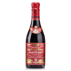 The image shows a bottle of Aceto Balsamico di Modena, which is balsamic vinegar from Modena, Italy. The bottle has a traditional shape with a red seal at the top. The label on the bottle is elaborately designed with a red and gold color scheme that gives it a premium look.

At the top of the label, there are two shields bearing coats of arms, and below them, the text "Gran Deposito Aceto Balsamico Giuseppe Giusti Modena" is prominently displayed. This indicates the product is from the Giuseppe Giusti estate, which is known for producing balsamic vinegar. The middle section of the label mentions "Dal 1605," suggesting the establishment of the estate in the year 1605.

The label also bears the Protected Geographical Indication (PGI) seal, which is the same as the seal you showed previously, indicating that the product is certified to have been produced in Modena with a quality and characteristics unique to that region. There are additional decorative elements and text on the label that likely provide more information about the product, its quality grade, and its heritage.