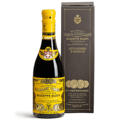 The image shows a bottle of Giuseppe Giusti Aceto Balsamico di Modena next to its packaging box. The bottle has a bright yellow cap and label, which features ornate gold and black designs, including the Giusti family crest. The label text indicates the prestigious Gran Deposito lineage, suggesting a high-quality vinegar aged according to traditional methods.

The box is a deep chocolate brown with gold lettering and accents, echoing the bottle's label design. It displays the Giuseppe Giusti logo at the top and the product name Aceto Balsamico di Modena, emphasizing the vinegar's origin and authenticity. The overall presentation is sophisticated and elegant, indicative of a premium product from a brand with a rich heritage.