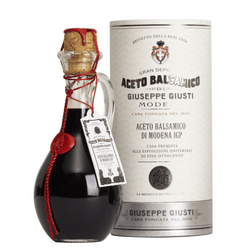 The image features a bottle of "Gran Deposito Aceto Balsamico di Modena IGP" by Giuseppe Giusti, accompanied by its cylindrical packaging. The bottle is rounded with a dark, almost black color, and is sealed with a gold-colored wax top, tied with a red decorative cord that has a seal attached. The packaging canister is white with red and black text, providing details about the product and its origin. This presentation underscores the traditional and premium nature of the vinegar, often used in gourmet cooking.