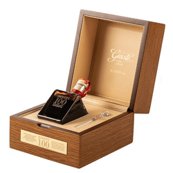 The image displays a very elegant and refined presentation of a balsamic vinegar bottle within a wooden gift box. Similar to the previous presentation, the bottle is uniquely shaped with a square base and an asymmetrical, tilted body, capped with a gold-colored lid. The label on the bottle proudly displays "100 anni", indicating it is a 100-year-old balsamic vinegar. The box is lined with a gold-colored interior, matching the luxurious theme, and has a golden plaque on the front that reads "Giusti Balsamico 100 anni". This setup highlights the extremely premium and exclusive nature of this vinegar, intended as a collector’s item or a significant gift.