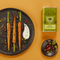 Green aluminum bottle of basilic olive oil with a tree in the middle with the brand name A l'Olivier on top on an orange background next to a beautiful black plate with carrots in it