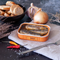 The image shows an open tin of sardines with an orange border on a fabric napkin, accompanied by a culinary setting. A knife rests beside the tin, indicating readiness for serving. The sardines are whole, their shiny skins and natural coloring suggesting a quality product.

Behind the tin, a bowl of sliced bread provides a rustic and hearty accompaniment, while an onion and a bulb of garlic add to the authenticity of the cooking ingredients, suggesting the potential for flavor enhancement. The scene also features a couple of red and yellow chili peppers, lending a hint of spice and vivid color that contrasts with the more subdued tones of the other elements.

Dried twigs and a sprinkling of coarse salt complete the composition, giving the image an earthy feel that complements the natural, simple quality of the sardines. The overall atmosphere of the setting is one of rustic simplicity, inviting the viewer to imagine enjoying a casual and flavorful meal.