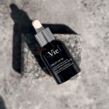 A dark glass serum bottle with a dropper cap, labeled "Sérum de Vie," resting on a textured stone surface.