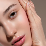 Close-up of a woman's face, showcasing smooth skin, soft lips, and a hand gently touching her cheek. Natural makeup and warm lighting.