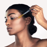 A woman applies a drop of serum to her cheek using a dropper, showcasing a skincare routine against a neutral background. A woman applies a drop of serum to her cheek using a dropper, showcasing a skincare routine against a neutral background.