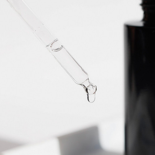 A clear dropper with a droplet of liquid at the tip, positioned near a dark bottle.