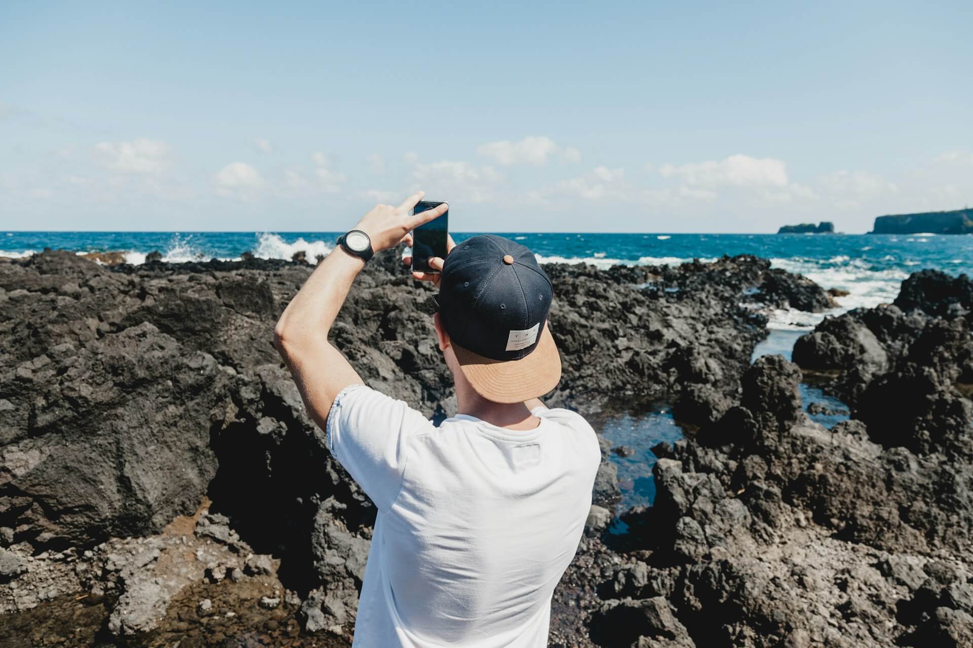 man taking photo of crashing ocean waves against rocks with smartphone