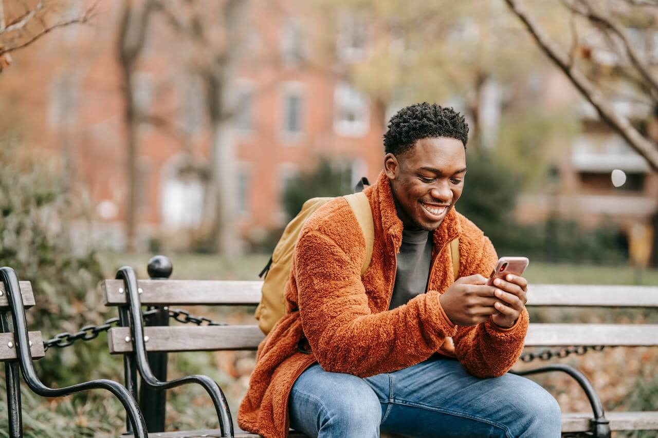 man sitting on a public bench and smiling at his smartphone