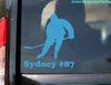 personalized ice hockey player vinyl decal featuring a skater holding a hockey stick with custom name Sydney and jersey number 87 beneath the silhouette, displayed on a red truck window, available in four sizes with mirror option