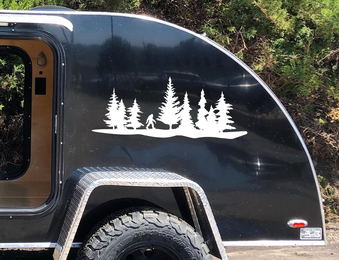 Bigfoot Sasquatch walking through pine forest tree line vinyl decal featuring evergreen trees and woodland landscape on RV camper storage compartment door