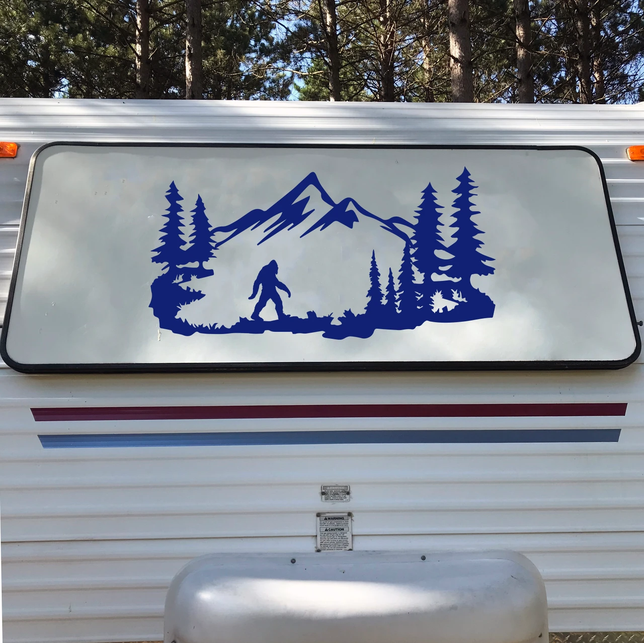 Bigfoot Sasquatch walking through mountain forest scene with evergreen pine trees and rugged peaks vinyl decal for RV camper and travel trailer