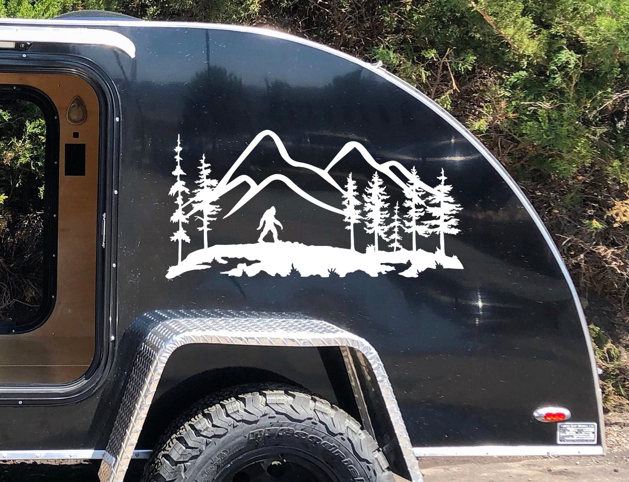 Bigfoot Sasquatch walking through mountain wilderness landscape with tall evergreen pine trees and rugged mountain peaks vinyl decal for RV camper and travel trailer