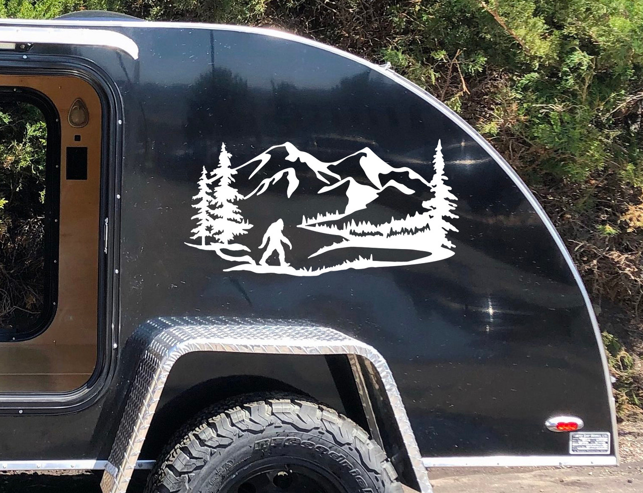 Bigfoot Sasquatch walking beside a wilderness lake with mountain peaks and evergreen pine trees scenic vinyl decal for RV camper and travel trailer