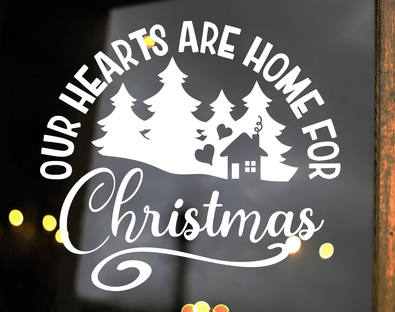 Our Hearts Are Home for Christmas vinyl window decal featuring evergreen pine trees, a small house, heart accents, arched lettering and decorative Christmas script