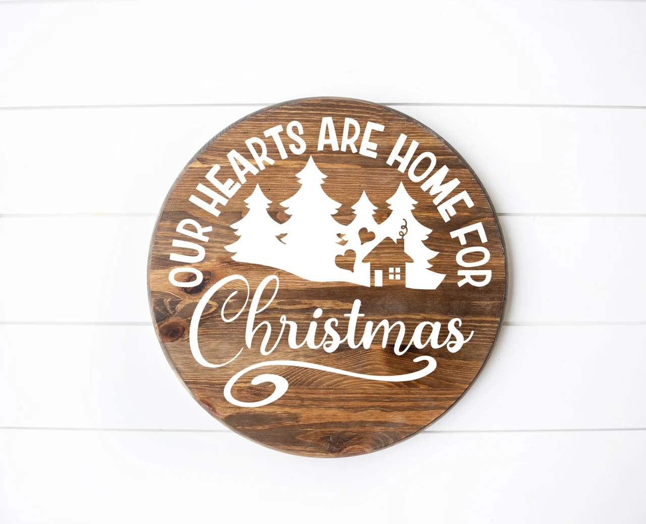 Our Hearts Are Home for Christmas vinyl window decal featuring evergreen pine trees, a small house, heart accents, arched lettering and decorative Christmas script