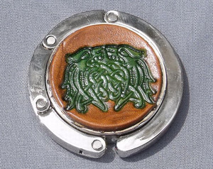 A purse hook with a leather medallion on it.  The medallion has a tan or light brown background with a green Celtic knot in the middle. The hook part is wrapped around the center and held closed with small magnets.