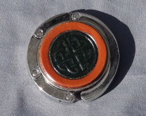 A purse hook with a leather medallion on it.  The medallion has a tan or light brown background with a Celtic knot that is dyed blue in the middle. The hook part is wrapped around the center and held closed with small magnets.