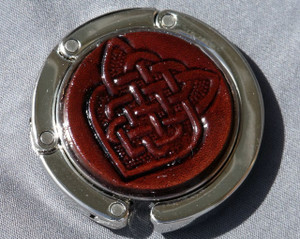 A purse hook with a leather medallion on it.  The medallion has a dark brown background with a celtic knot in the middle. The hook part is wrapped around the center and held closed with small magnets.