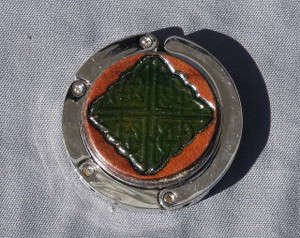A purse hook with a leather medallion on it.  The medallion has a tan or light brown background with a green celtic knot in the middle. The hook part is wrapped around the center and held closed with small magnets.