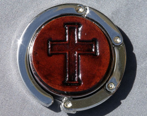 A purse hook with a leather medallion on it.  The medallion has a dark brown background with a cross in the middle. The hook part is wrapped around the center and held closed with small magnets.