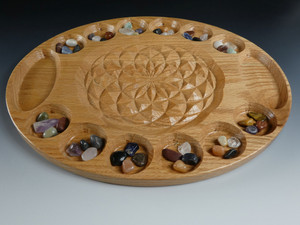 The large deluxe mancala board in oak with the flower of life in the center and beautiful stones in their starting positions.