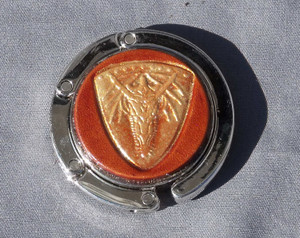 A purse hook with a leather medallion on it.  The medallion has a tan or light brown background with a gold dragon shield in the middle. The hook part is wrapped around the center and held closed with small magnets.