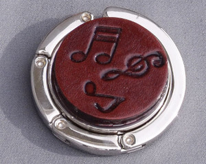 A purse hook with a leather medallion on it.  The medallion has a dark mahogany background with musical notes and symbols randomly placed in the middle. The hook part is wrapped around the center and held closed with small magnets.