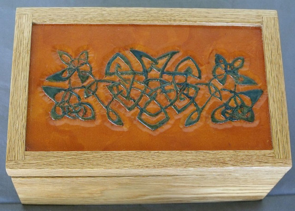 A rectangular red oak keepsake box showcasing a prominent top inset paneled with genuine leather, which is hand-carved with a classic Celtic knot design and dyed deep blue.